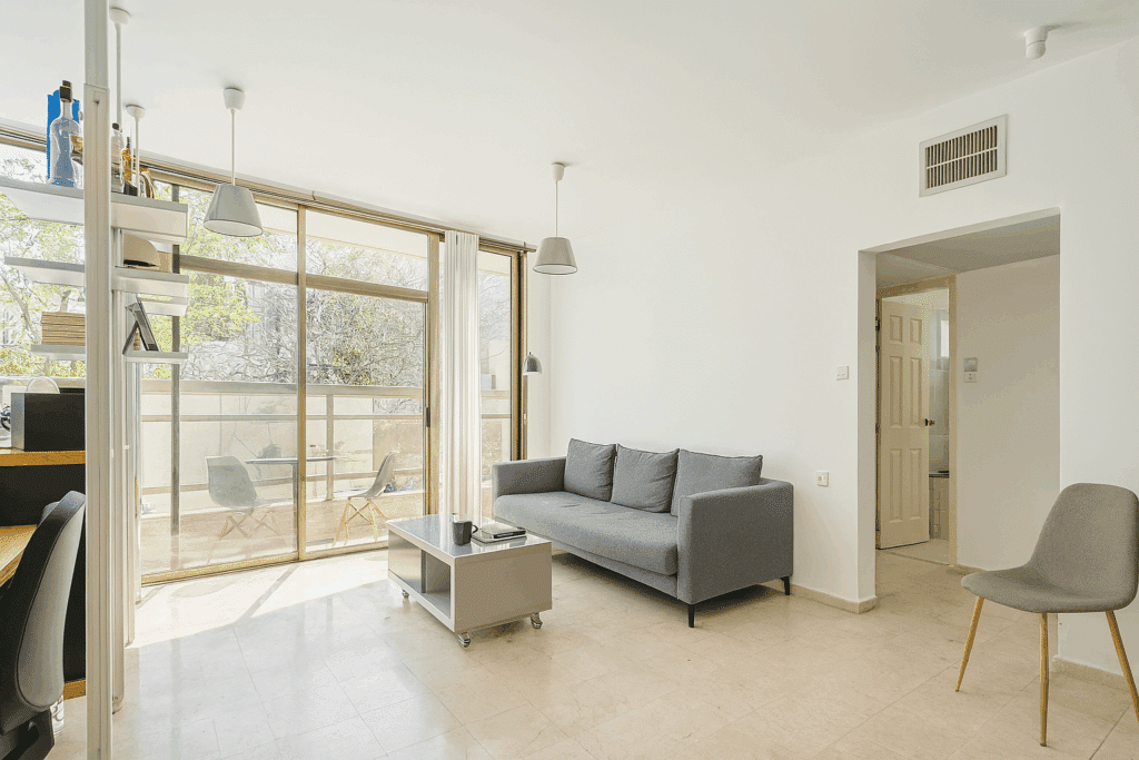 Bright living room with modern gray sofa and large windows opening to a green balcony – apartment for sale in Tel Aviv
